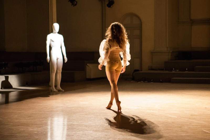 A woman in a billowy blouse faces a mannequin as she stands on her tip toes